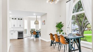 A spacious kitchen with white cabinets, a dining area with a table set for four, and a large window with a view of a tree and a parked car. - Newly Renovated, Spacious & Modern | THE (Rockwall)