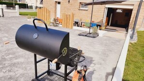 Una parrilla negra con ruedas, un tanque de gas y una bolsa de carbón sobre un patio de concreto.