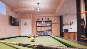 Una mesa de billar con bolas y tacos, una estantería con varios objetos, y una zona de cocina con un microondas.