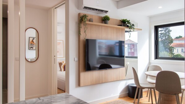 Uma sala de estar moderna com uma televisão de tela plana grande fixada em uma parede de madeira, um espaço de jantar com mesa e cadeiras, e vista para o exterior por uma grande janela.