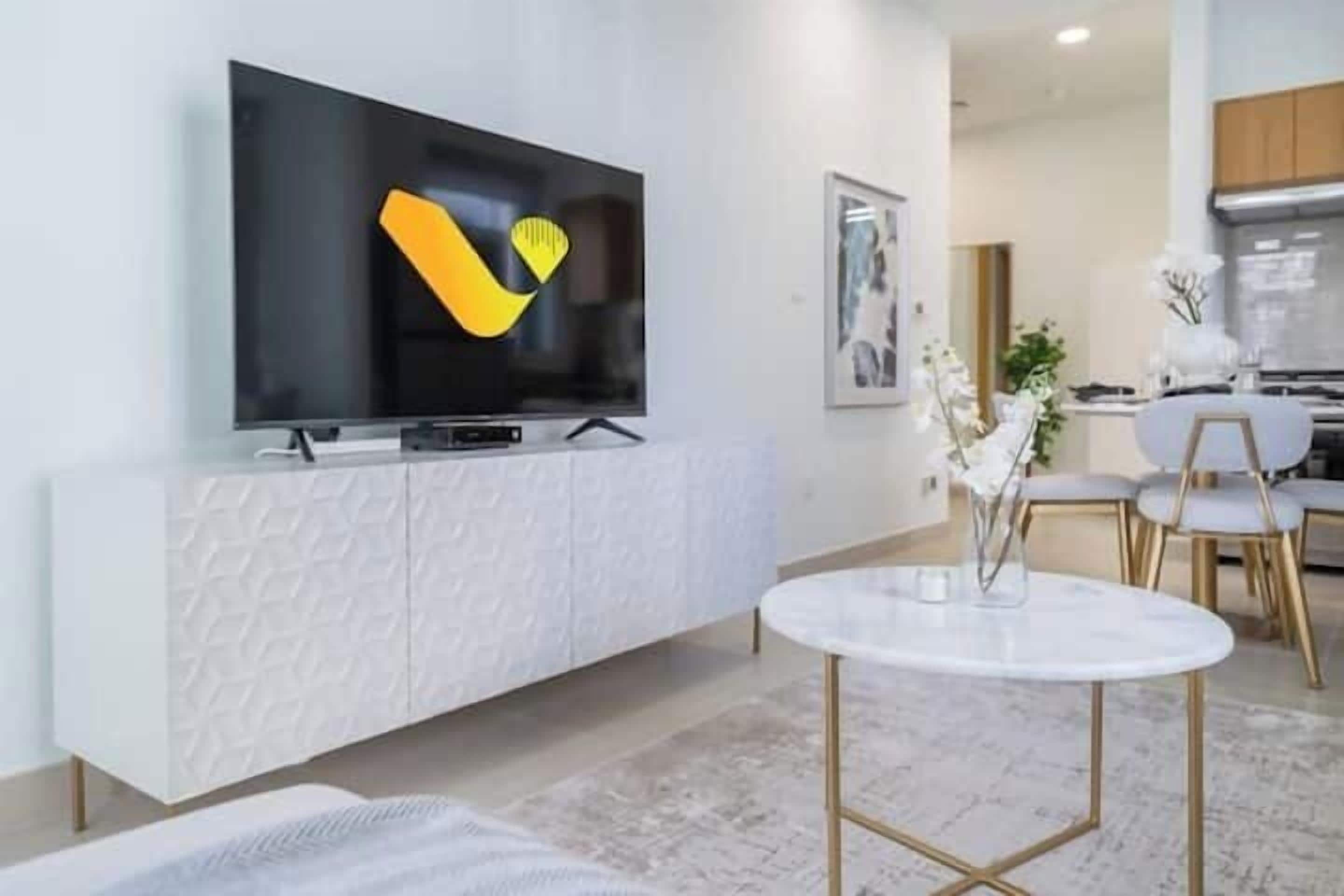 Una sala moderna con un televisor de pantalla plana montado sobre un mueble blanco texturizado, una mesa redonda con patas doradas y un comedor con sillas blancas y una pequeña mesa.