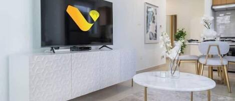 Una sala moderna con un televisor de pantalla plana montado sobre un mueble blanco texturizado, una mesa redonda con patas doradas y un comedor con sillas blancas y una pequeña mesa.