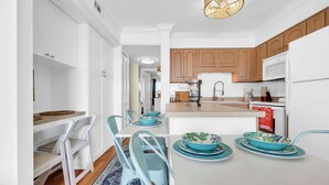 A modern kitchen with white cabinetry, a central island with a sink, and a dining area with blue chairs and plates. - Beach Escape Top Location w Pool (North Myrtle Beach)