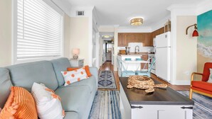 A modern living room with a grey sofa, a coffee table, and a kitchen in the background. - Beach Escape Top Location w Pool (North Myrtle Beach)