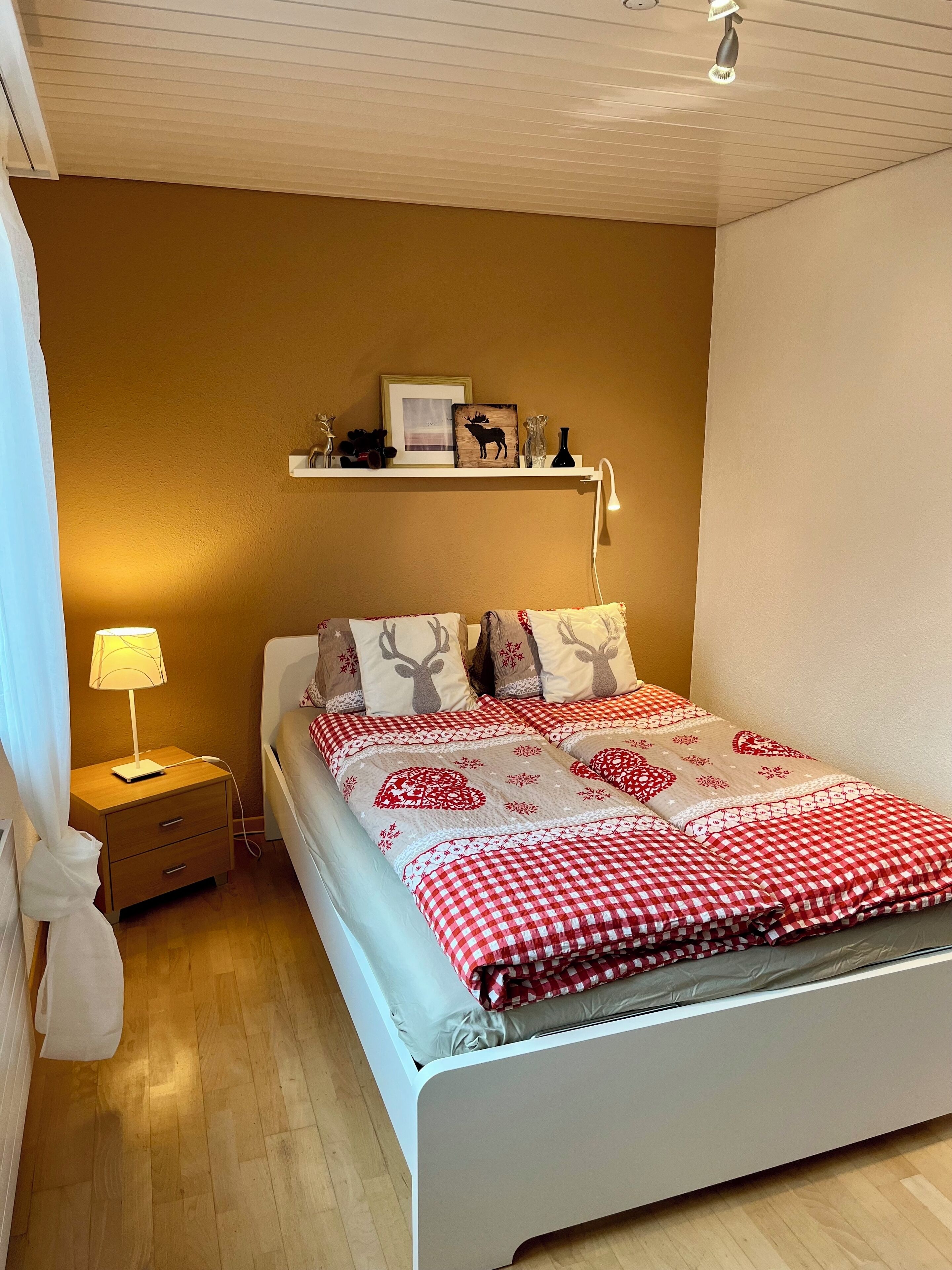 A neatly arranged bedroom with a bed, a bedside table with a lamp, a shelf with decorative items, and a wall-mounted light fixture.