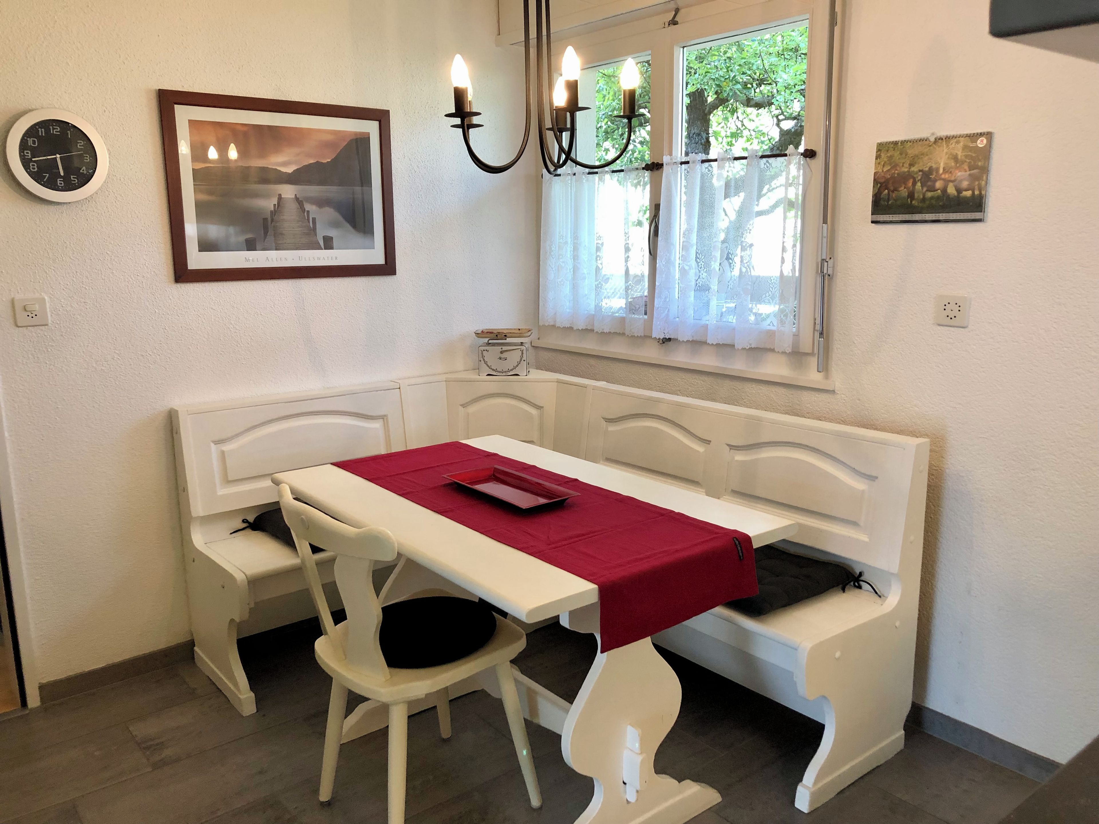 Een kamer met een witte bank, een tafel met een rode tafelkleed, een klok, een ingelijst beeld en een raam met kantgordijnen.