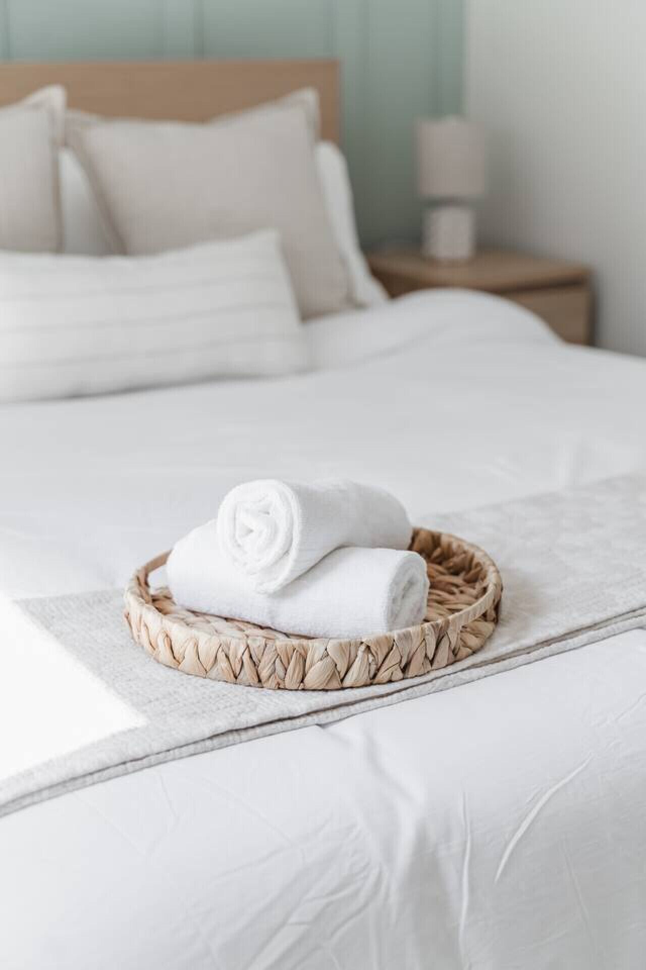 Un lit bien fait, avec du linge de lit blanc et un plateau en osier contenant deux serviettes enroulées.
