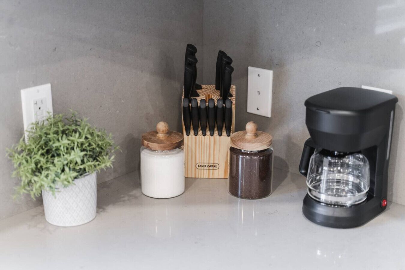 A kitchen counter with a coffee maker, two jars, a knife block, and a potted plant.