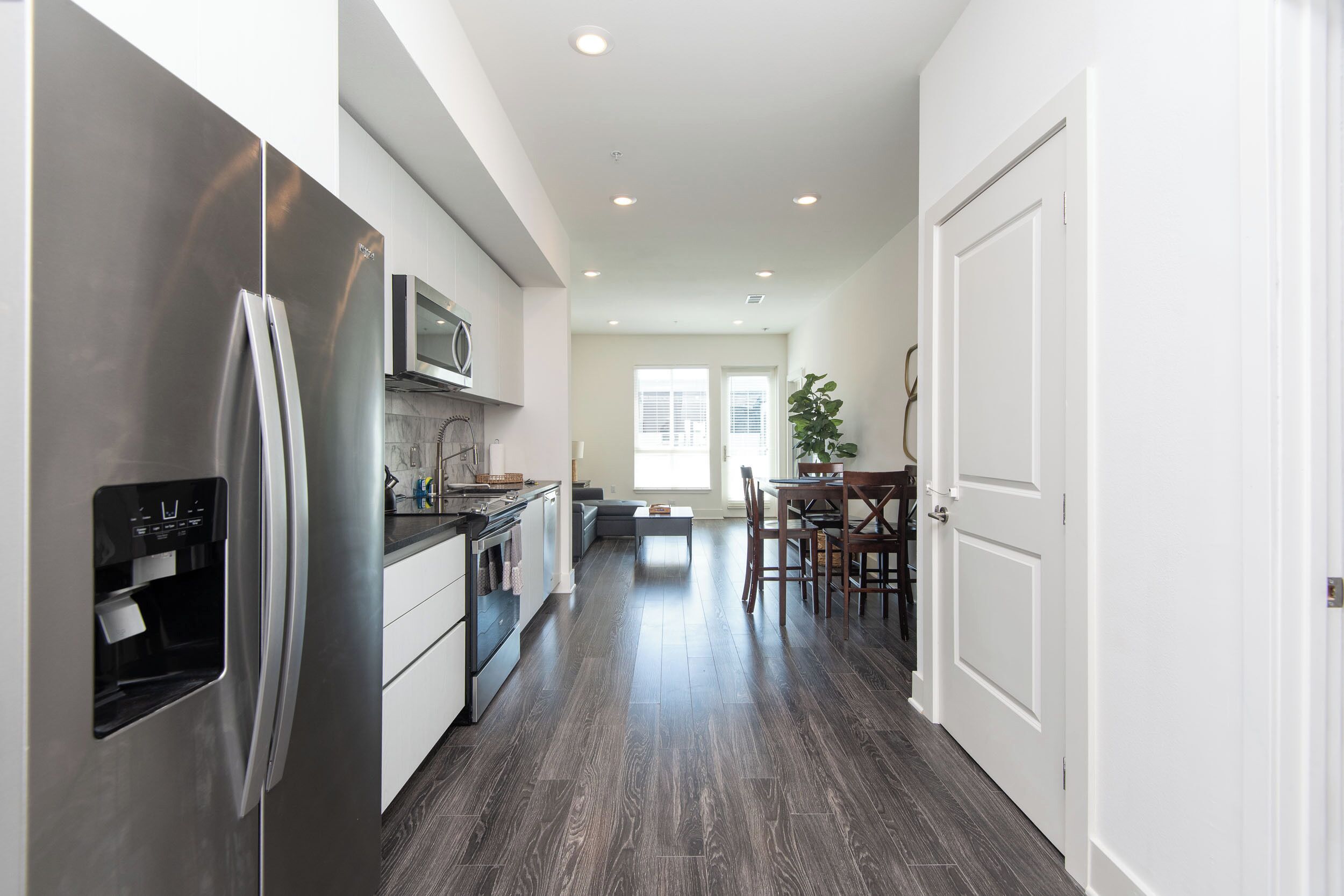 A modern kitchen with stainless steel appliances, a dining area with a table and chairs, and a hallway leading to a door.