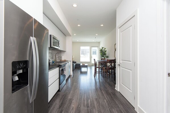 A modern kitchen with stainless steel appliances, a dining area with a table and chairs, and a hallway leading to a door.