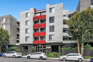 A multi-story building with a red balcony on the second floor, a visible entrance, and several parked cars. - 2BR Hollywood Apt w/ Rooftop Pool & Stunning Views (Los Angeles)
