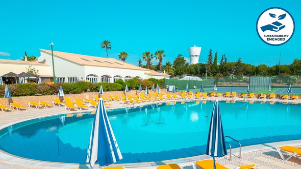 Una piscina con sillas de descanso amarillas y sombrillas azules, rodeada de vegetación y un edificio de fachada blanca.