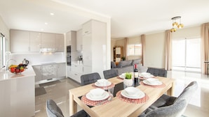 Una cocina moderna con armarios blancos, un comedor con una mesa de madera dispuesta para cuatro personas y una sala de estar con un sofá y una televisión.
