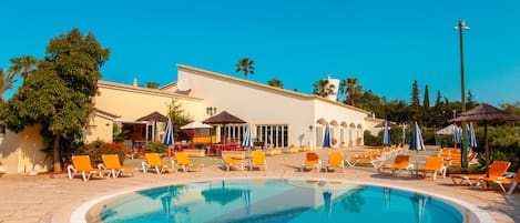 A pool area with lounge chairs and umbrellas, a building with a tiled roof, and palm trees.