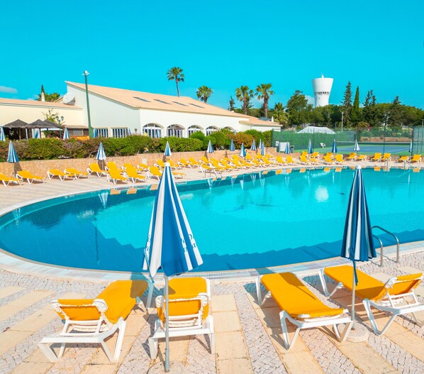 A swimming pool with yellow lounge chairs and blue umbrellas, surrounded by greenery and a building with a white facade.