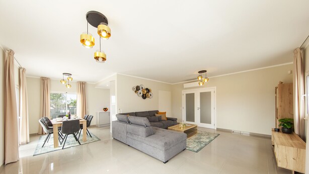 A spacious living area with a grey sectional sofa, a dining table with chairs, and a coffee table. There are pendant lights above the sofa and a wall-mounted light fixture. - Villa Oasis Gold (Portimão)