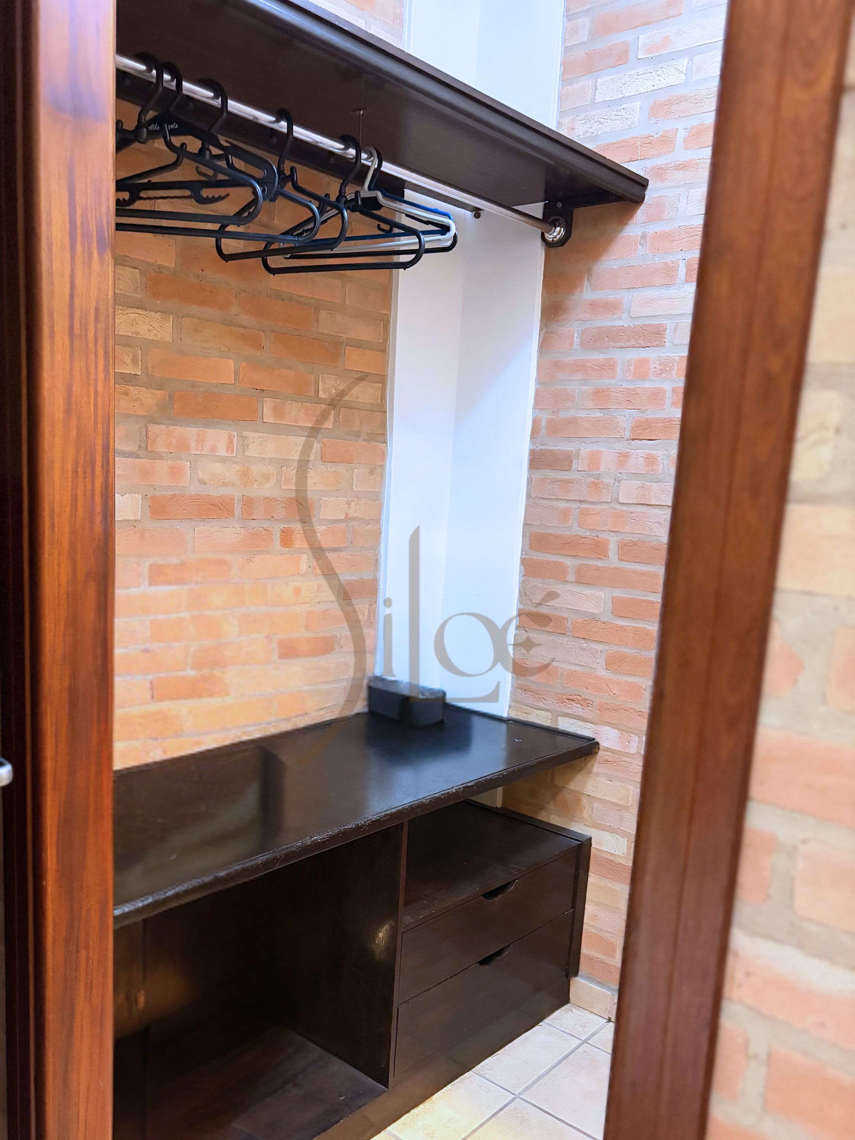 A wooden wardrobe with a hanging rack and a white vertical object, possibly a towel or a sign, against a brick wall.