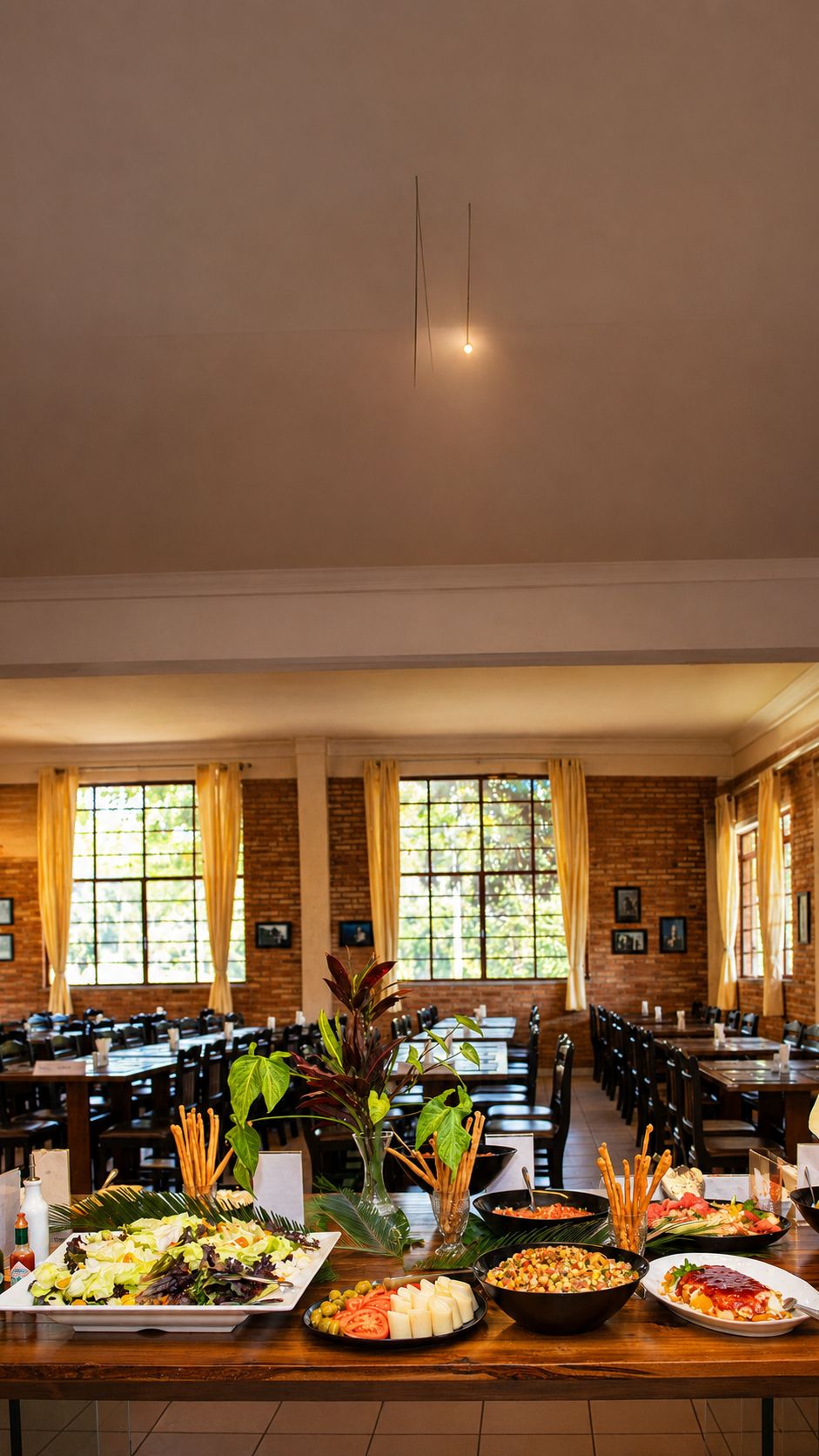 Uma mesa de buffet montada em área de refeições, com várias mesas e cadeiras, janelas amplas com cortinas e uma parede de tijolos.