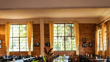 Uma mesa de buffet montada em área de refeições, com várias mesas e cadeiras, janelas amplas com cortinas e uma parede de tijolos.