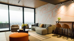 A modern living room with a sofa, ottoman, and bar stools. There is a large window providing a view of the city.
