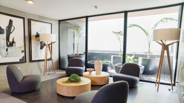 A modern living room with a sofa, armchairs, a coffee table, and floor lamps. There are large windows with a view of the city and plants.