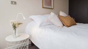A neatly made bed with white bedding and a brown pillow, a white bedside table with a lamp and a vase of flowers, and a framed picture on the wall.