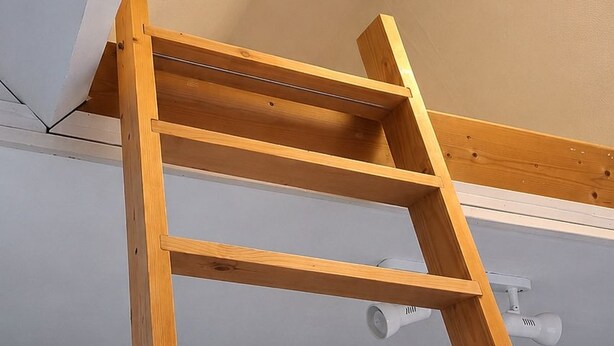 A wooden ladder in a room with a sloped ceiling and a ceiling light fixture. - Mixed-gender shared room – Double bed on a loft (Seraing)