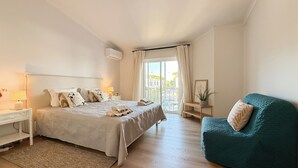 Une chambre à coucher comprenant un lit, des tables de chevet, une chaise, une fenêtre avec des rideaux et une vue sur l’extérieur.