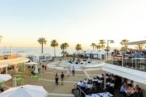 A rooftop bar with outdoor seating overlooking the sea, palm trees, and a clear sky.
