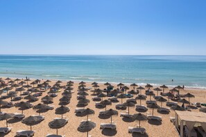 A beach with numerous sun loungers and thatched umbrellas, a clear sky, and the ocean in the background.