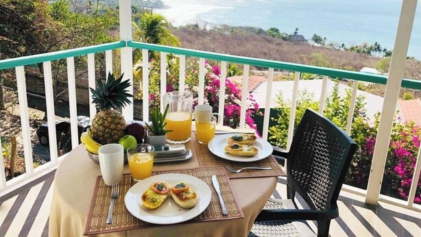A breakfast table on a balcony with a view of the ocean, a pineapple, and a pitcher of orange juice.