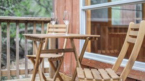 Een houten terras met een tafel en stoelen, twee lege wijnglazen en uitzicht op bomen door een raam.
