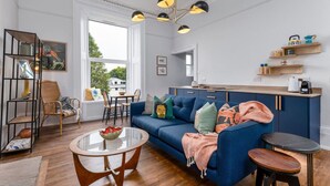 A modern living room with a blue sofa, a glass coffee table, and a dining area with a wooden table and chairs. - Stirling Retreat - Quirky Décor, Fireplace & Charm (Stirling)