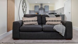 A modern living room with a grey sofa, patterned throw pillows, a black floor lamp, and a kitchen in the background. - Stirling's Hidden & Tranquil Studio Apartment (Stirling)