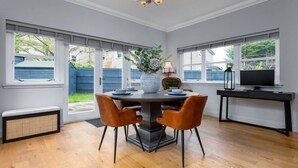 A dining area with a round table, two chairs, a vase with flowers, a television on a stand, and a window with blinds.