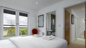 A modern bedroom with a large bed, a view through the window, a mirror, and a bathroom visible through an open door. - The Stirling Penthouse - Free Parking (Stirling)