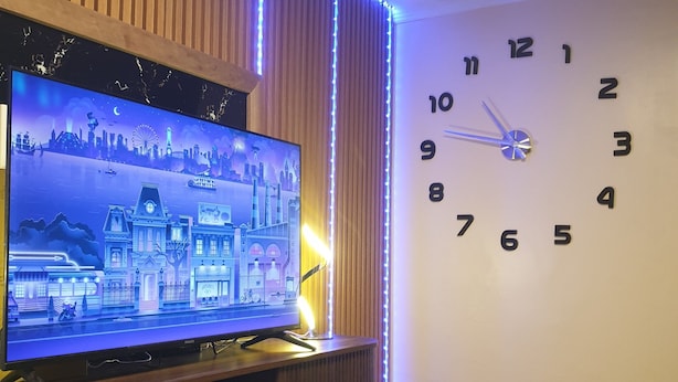 A living room with a flat-screen TV on a wooden stand, a wall clock, and a seating area with a table and chairs.