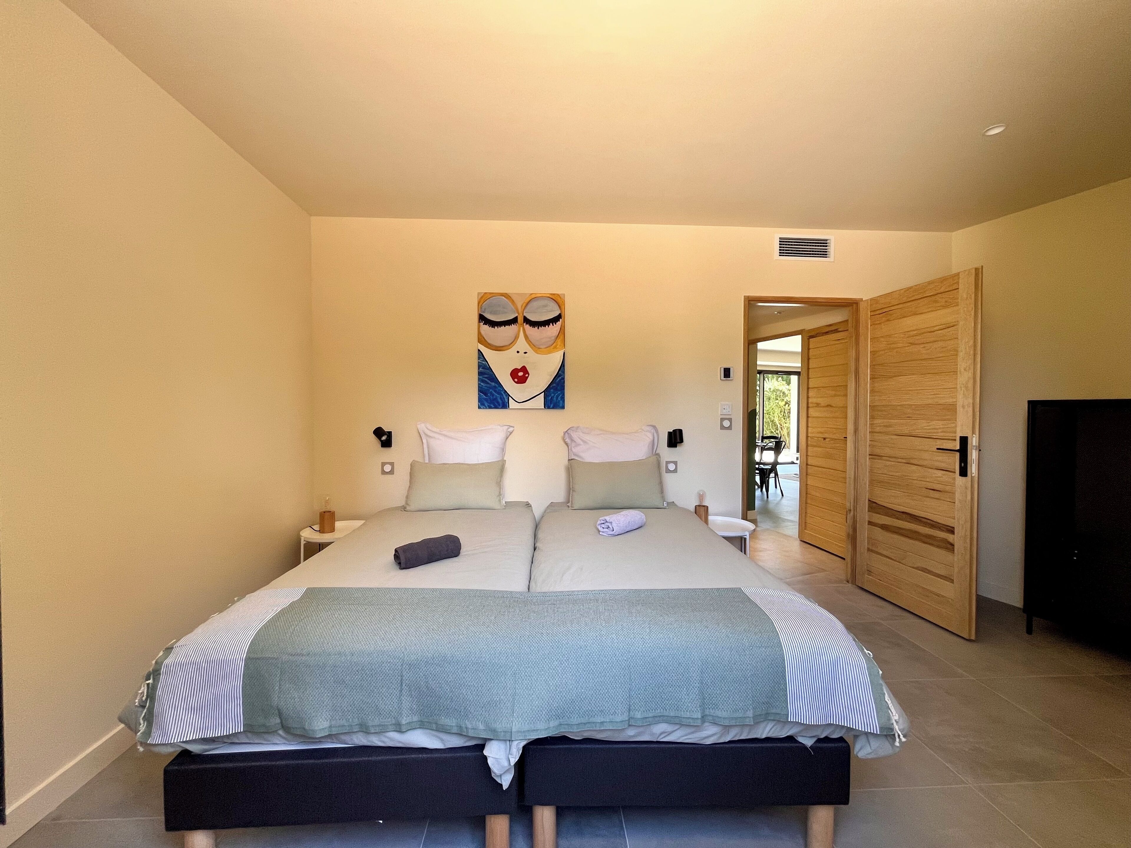 Ein Schlafzimmer mit einem großen Bett, zwei Nachttischen, einem an der Wand befestigten Kunstwerk und einer Holztür, die zu einem Außenbereich führt.