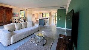 Een moderne woonkamer met een bank, een glazen salontafel en een televisie. In de achtergrond is een keuken met een eethoek te zien.