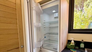 Een kleine keuken met een inbouwkoelkast, een raam met uitzicht op groen, en een aanrecht met schoonmaakmiddelen.