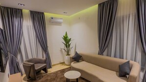 A living room with a beige sofa, a brown rug, a small round table, a grey armchair, and a potted plant. - The Corniche Hotel and Apartment (Lekki)