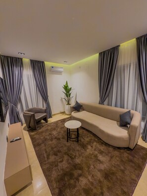 A living room with a beige sofa, a brown rug, a small round table, a grey armchair, and a potted plant. - The Corniche Hotel and Apartment (Lagos)