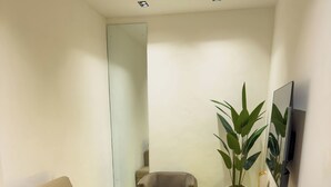 A small, modern living room with a sofa, a chair, a small round table, a potted plant, and a flat-screen TV mounted on the wall. - The Corniche Hotel and Apartment (Lekki)