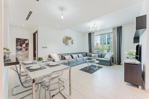 A modern living room with a grey sofa, a dining table with chairs, a TV on a stand, and a large window with curtains. - PVH - Serene 2BR - Sobha Hartland Greens (Dubai)
