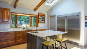 Une cuisine moderne avec des meubles en bois, un grand îlot central doté de tabourets, un réfrigérateur en acier inoxydable et une lucarne au-dessus de l’évier.