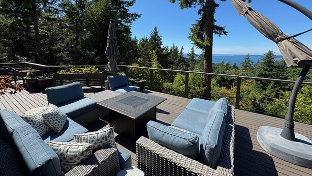 Une terrasse aménagée avec du mobilier d’extérieur, notamment des canapés et une table, offrant une vue sur une zone boisée et l’océan.