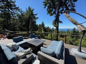 En terrasse med udendørs møbler, herunder sofaer og et bord, med udsigt over et skovområde og havet.