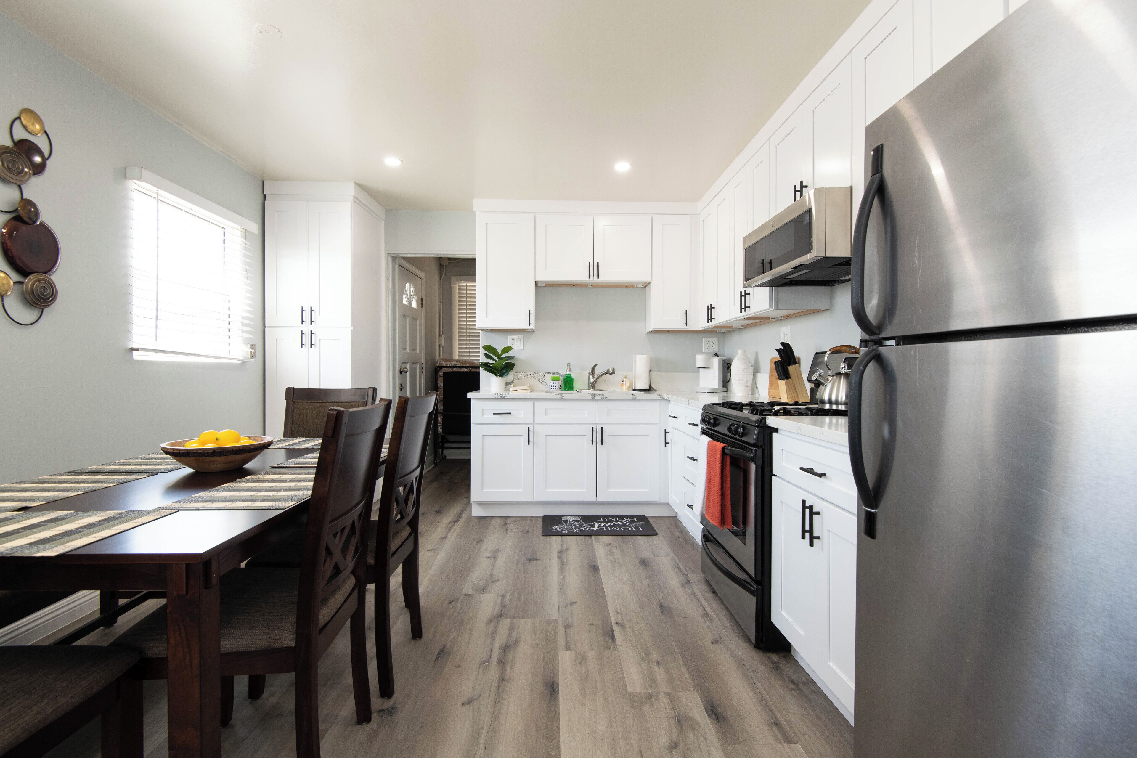 Una cocina moderna con armarios blancos, un refrigerador de acero inoxidable y un comedor con mesa y sillas de madera.