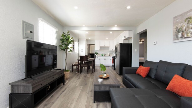 A modern living room with a flat-screen TV, a dark-colored sofa with orange cushions, a coffee table, and a dining area with a wooden table and chairs.