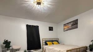 Un dormitorio con cama, mesita de noche, silla, una pequeña mesa, planta en maceta y lámpara en el techo.